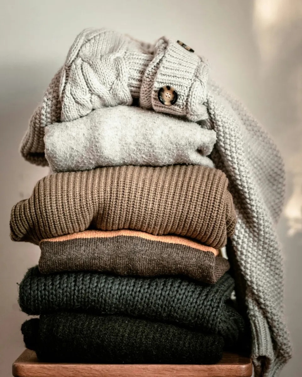 A stack of folded knitwear in cream, grey, brown and green on a wooden stool, in warm window light