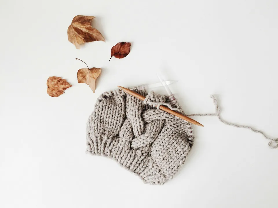 Grey cable knitting on a wooden needle beside autumn leaves